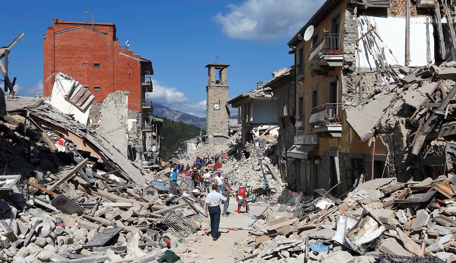 Photograph of ruined buildings and piles of rubble with an intact clock tower still standing in the background. People walk among the rubble.