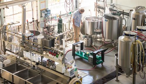 A man stands over a large pot surrounded by tanks, tubes and other equipment. 