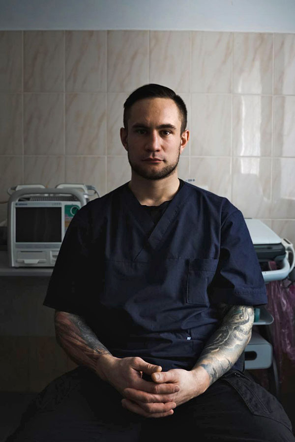 A serious young man in dark blue scrubs, his tattooed arms folded in front of him, in a hospital room.