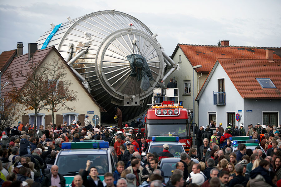 Photo shows the transport of the massive, metallic KATRIN spectrometer down a narrow street, overhanging the houses on either side, in the center of Leopoldshafen, Germany, with people gathered for the event.
