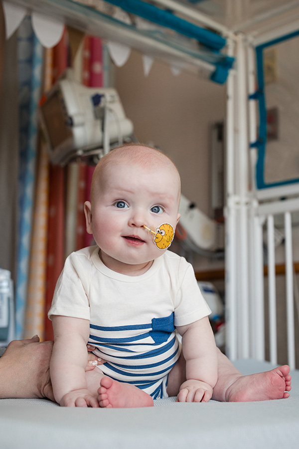 A young baby sits in a crib with a tube in his nose affixed with a Big Bird sticker on his cheek.