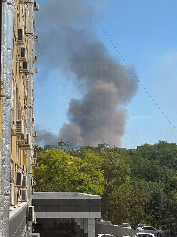 A large dark cloud of dust and smoke fills the sky behind trees and a building.