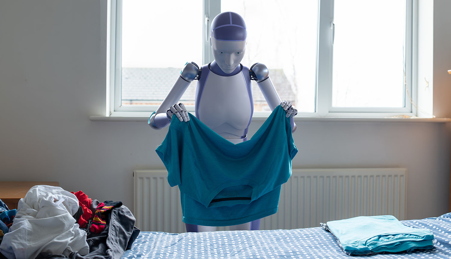 A humanoid robot stands in front of a window, folding a large T shirt.